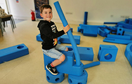 A smiling boy rides a creation which he has made from bright blue building blocks which resembles a steam train engine or tractor
